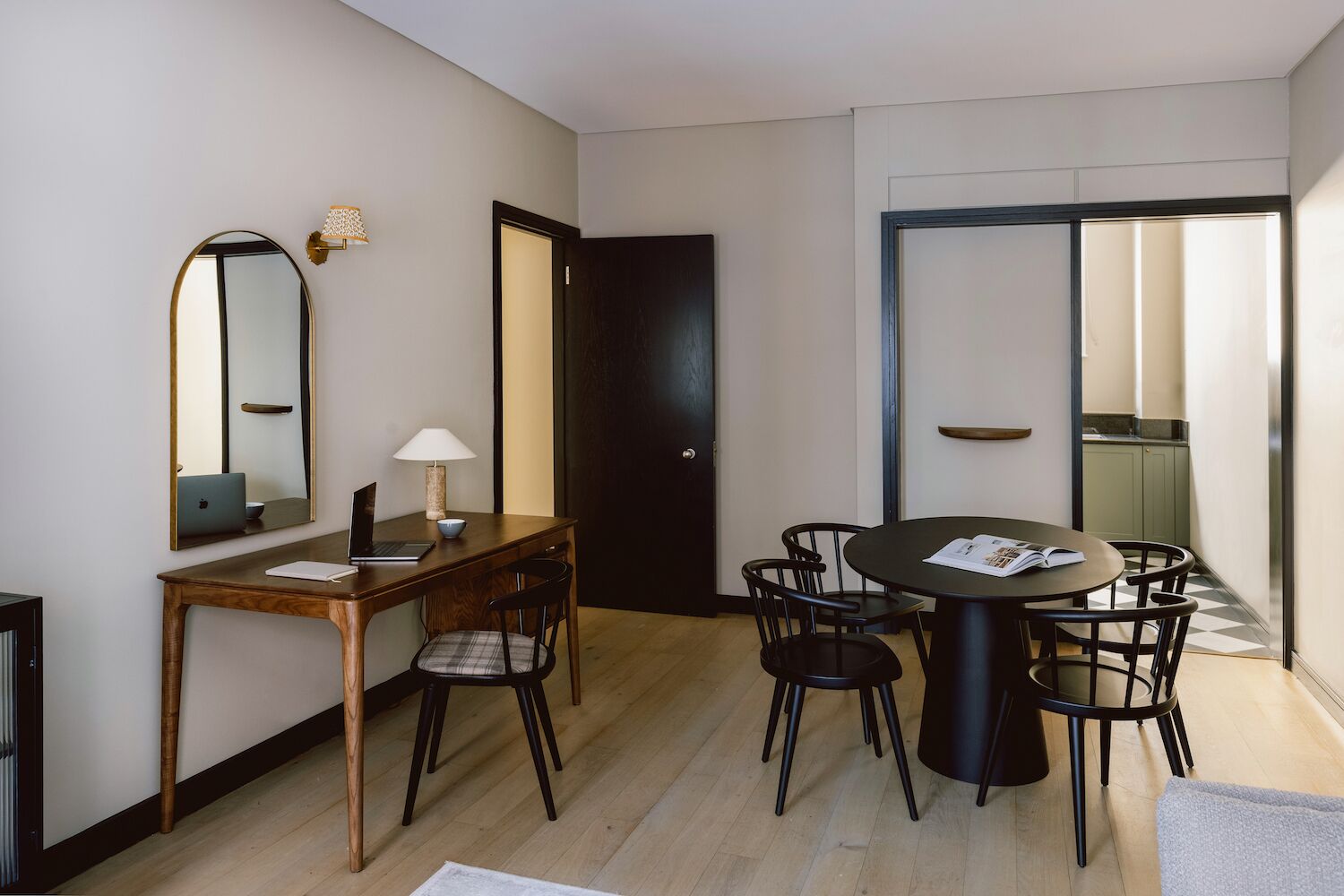 Una habitación moderna y bien iluminada, con un escritorio de madera, dos sillas, una mesa redonda con una revista, una computadora portátil y una lámpara.
