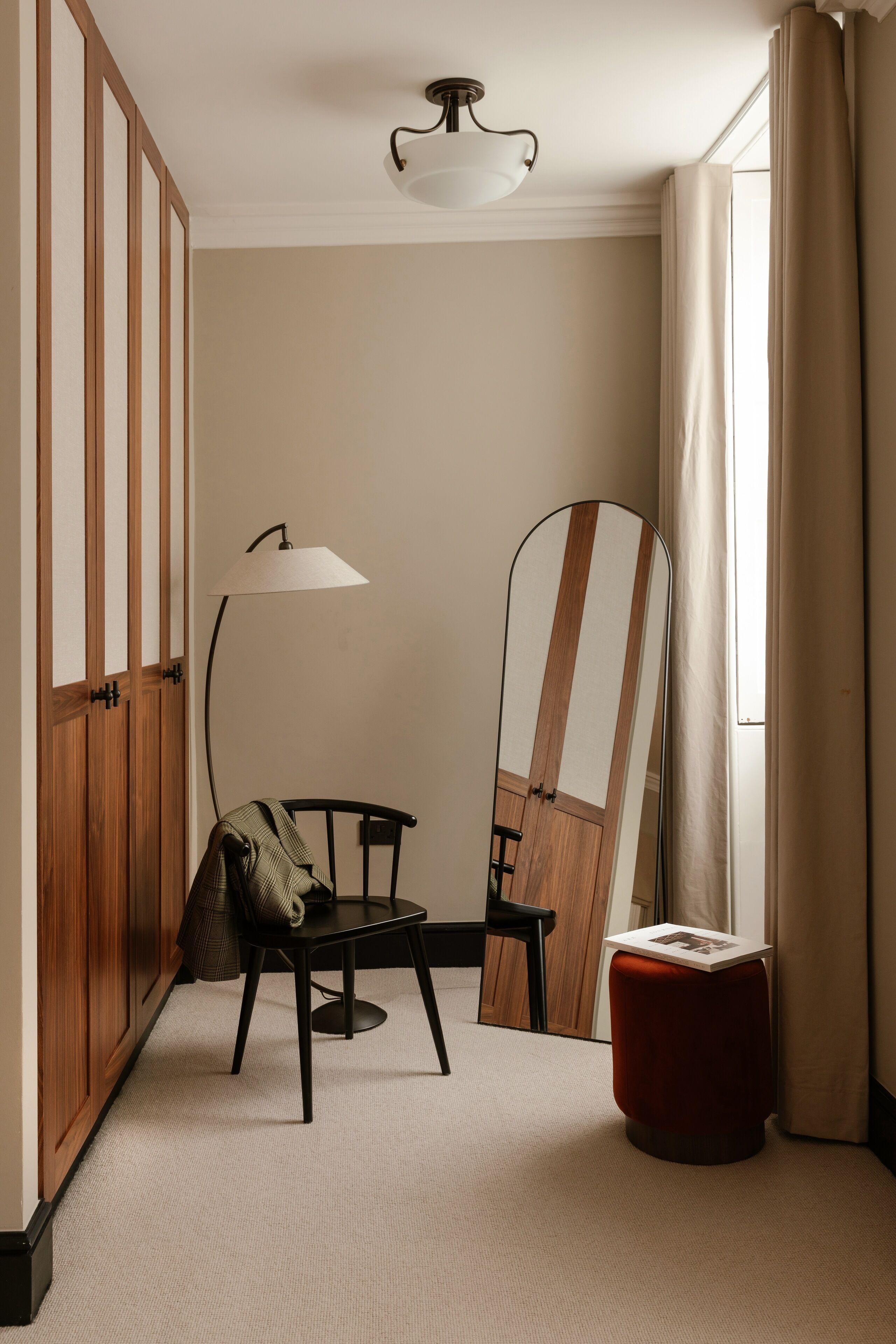 Una habitación con un armario de madera, una mesa redonda con una silla, una lámpara de pie, un espejo y un otoman rojo.