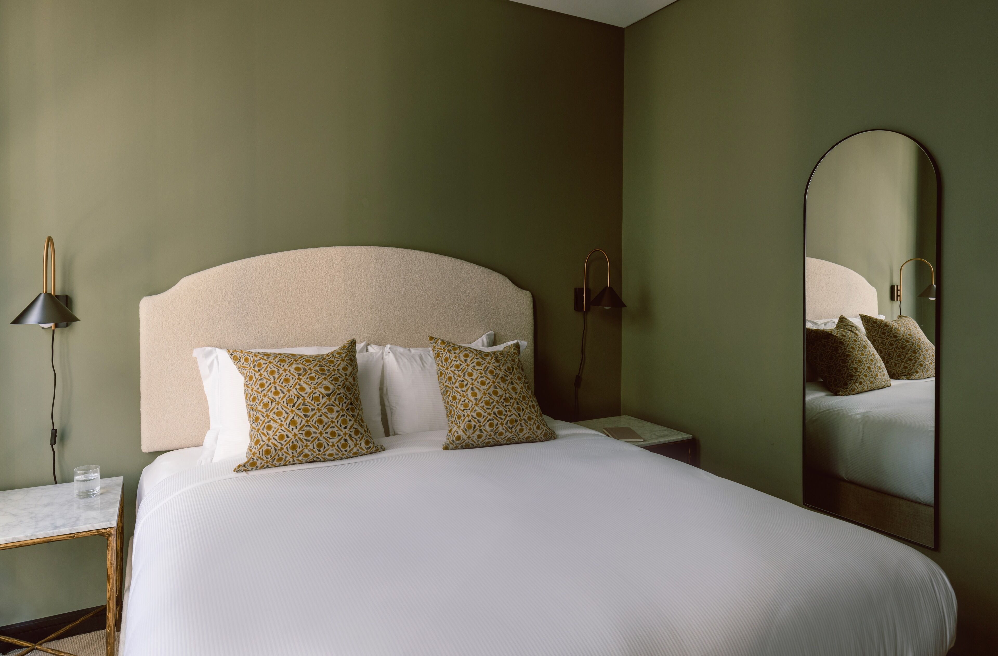 A neatly made bed with patterned pillows, a bedside table with a lamp, and a large mirror on the wall.