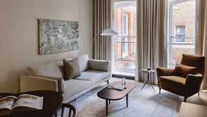 Ein modernes Wohnzimmer mit einer Couch, einem Sessel, einem Couchtisch und einem kleinen Beistelltisch. Ein großes Fenster mit Vorhängen bietet Blick auf ein Backsteinhaus.
