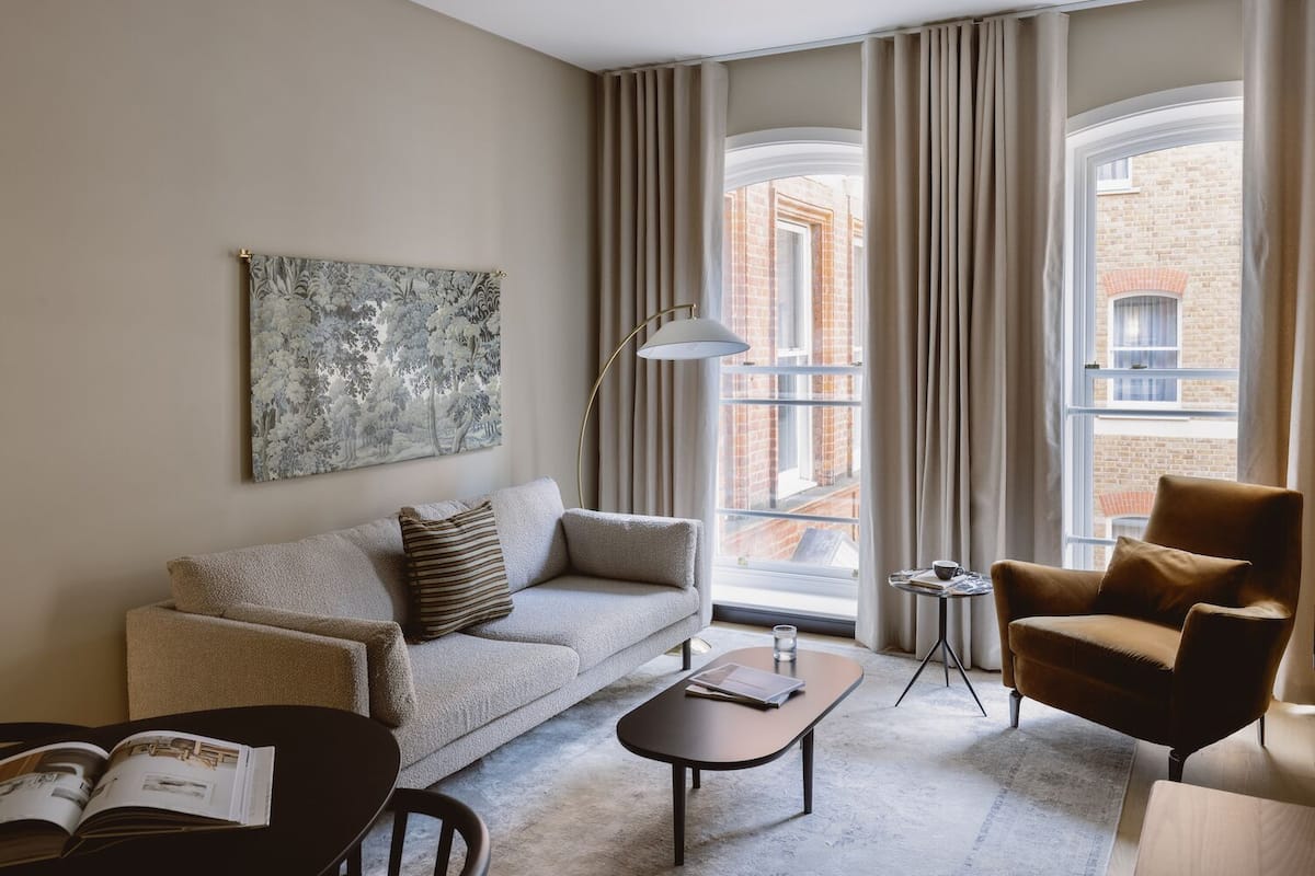 A modern living room with a sofa, armchair, coffee table, and a small side table. A large window with curtains provides a view of a brick building.
