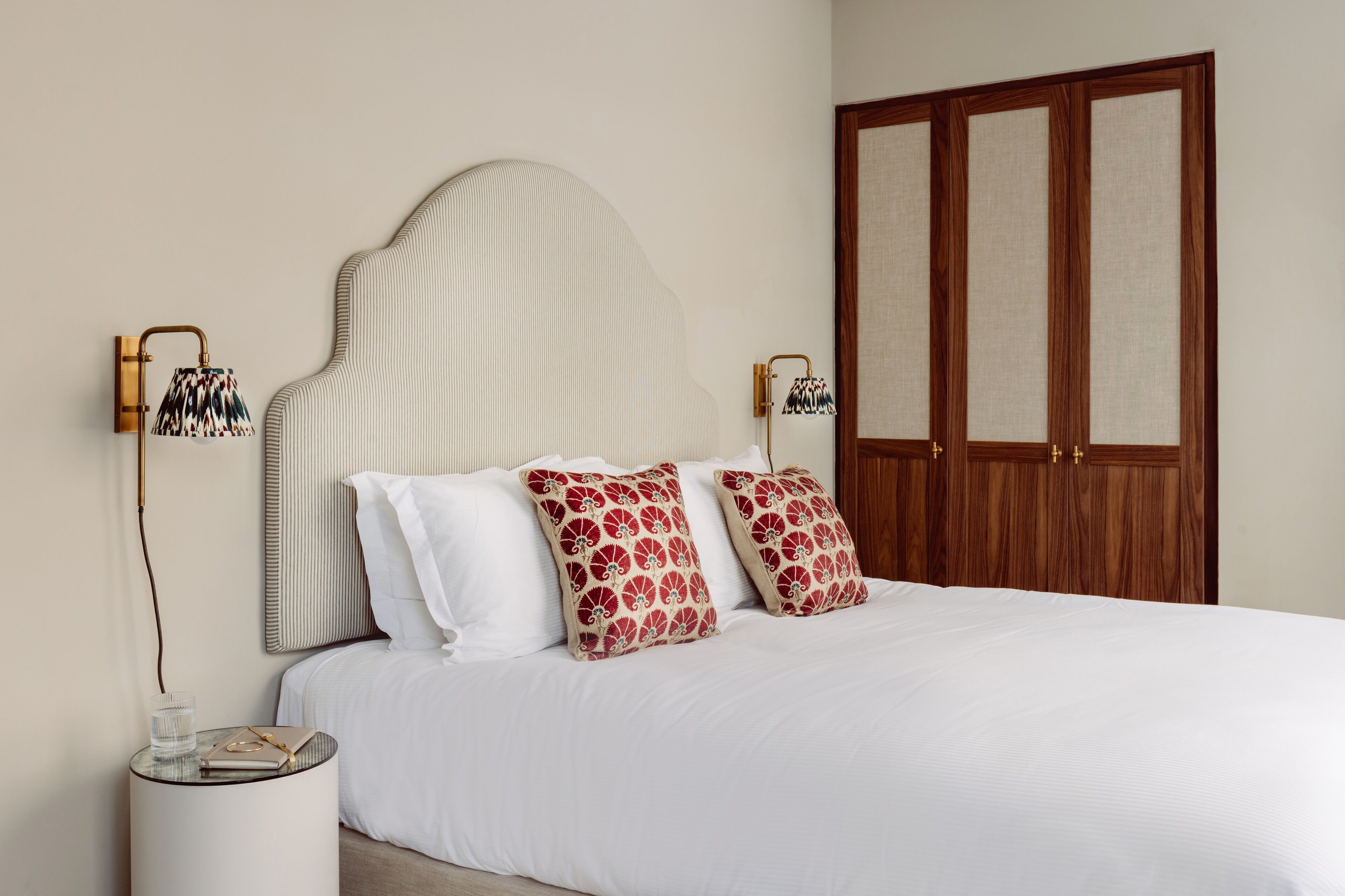 Een netjes opgemaakt bed met witte beddengoed en gestreepte kussens, een nachtkastje met een lamp en een houten garderobe.