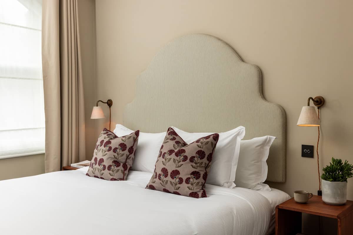 A neatly made bed with white linens and two decorative pillows, a bedside table with a lamp, and a potted plant on the nightstand.