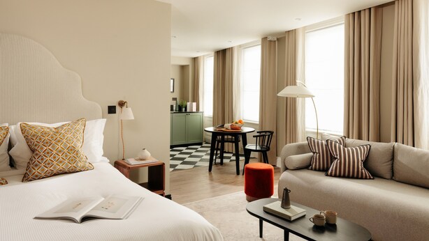 Ein Hotelzimmer mit Bett, einer Couch, einem Esstisch mit Stühlen, einer Küchenzeile und großen Fenstern mit Vorhängen.