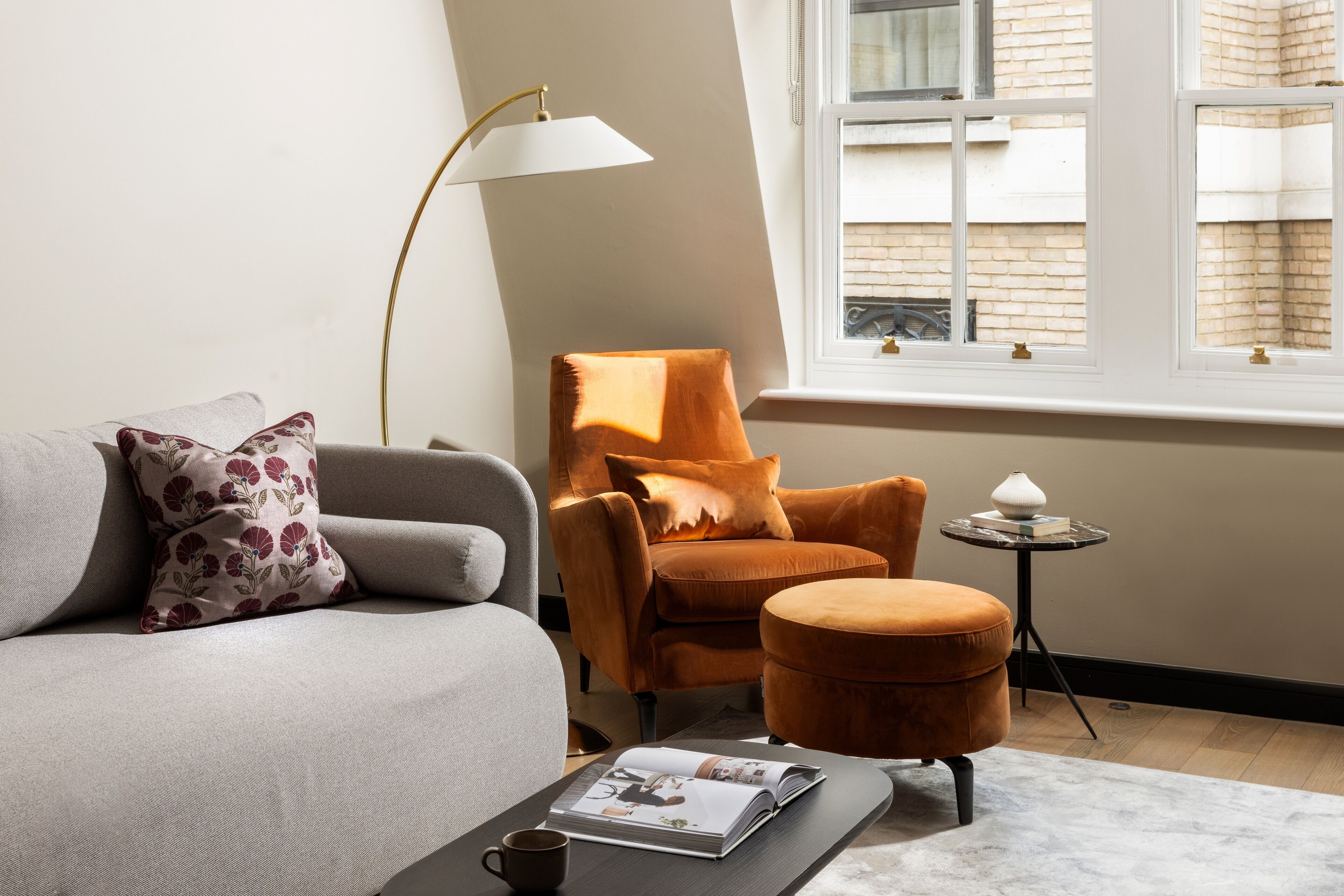Een gezellige woonkamer met een grijze bank, een oranje fauteuil, een klein rond voetenbankje, een vloerlamp en een tijdschrift op een salontafel.