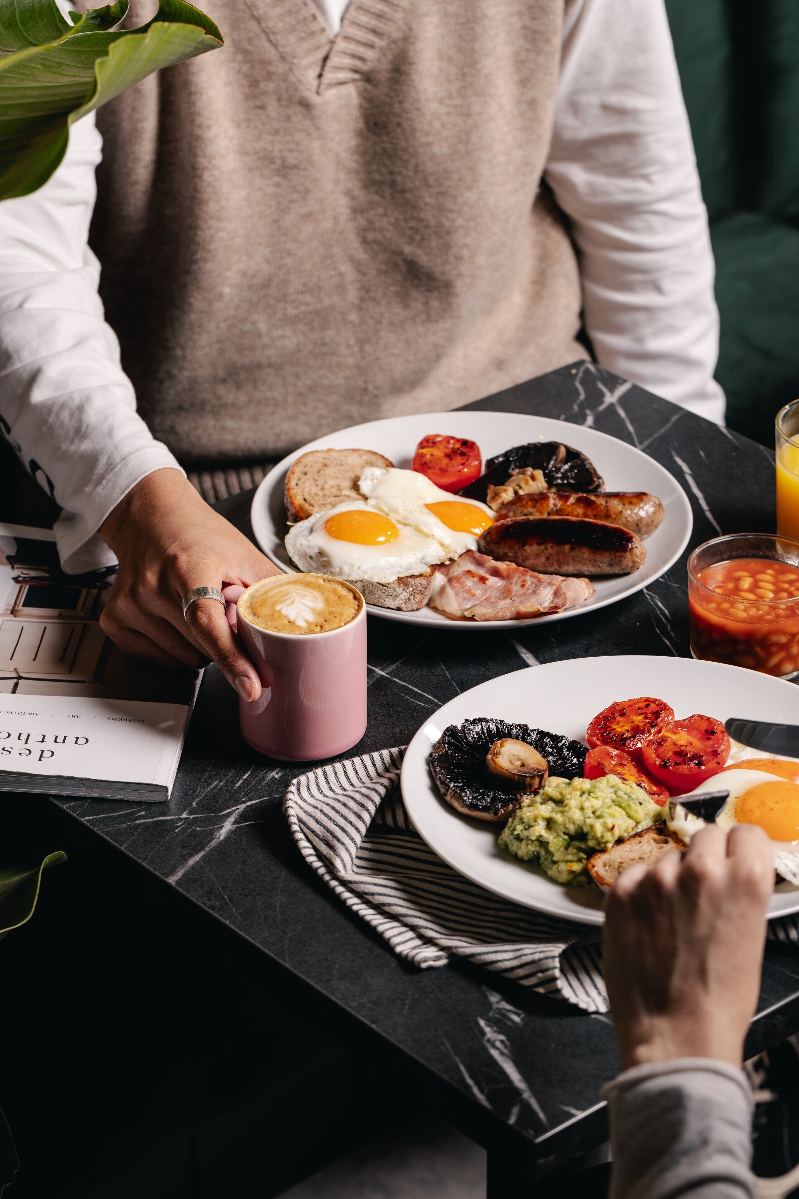 A breakfast setting with plates of eggs, sausages, toast, and avocado on a marble table.