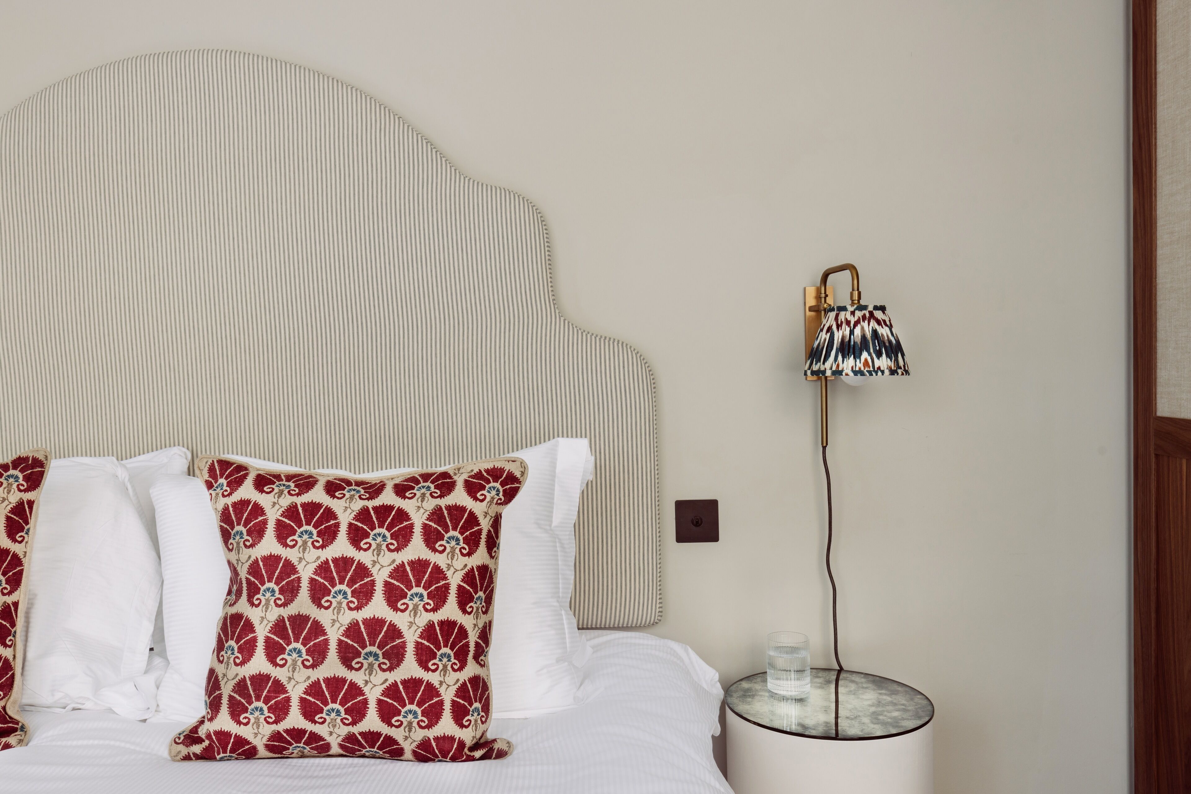 Un letto rifatto con cuscini bianchi e rossi a motivi decorativi, una testiera a righe e un comodino con una lampada e un recipiente di vetro.
