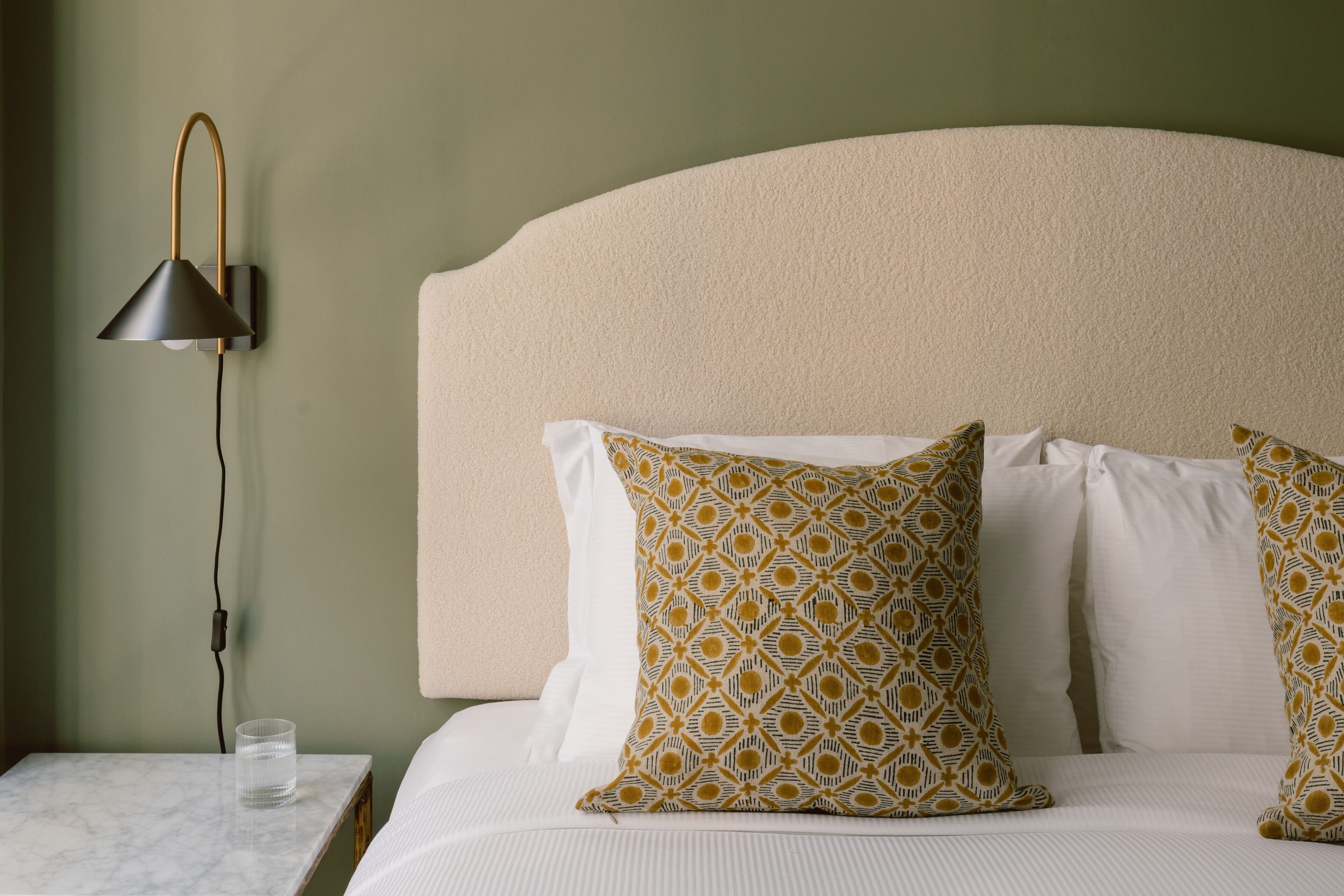 A neatly made bed with patterned pillows, a bedside lamp, and a glass of water on a marble nightstand.