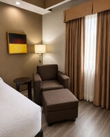 A hotel room with a bed, a sofa, an ottoman, a lamp, a painting, and a window with curtains.