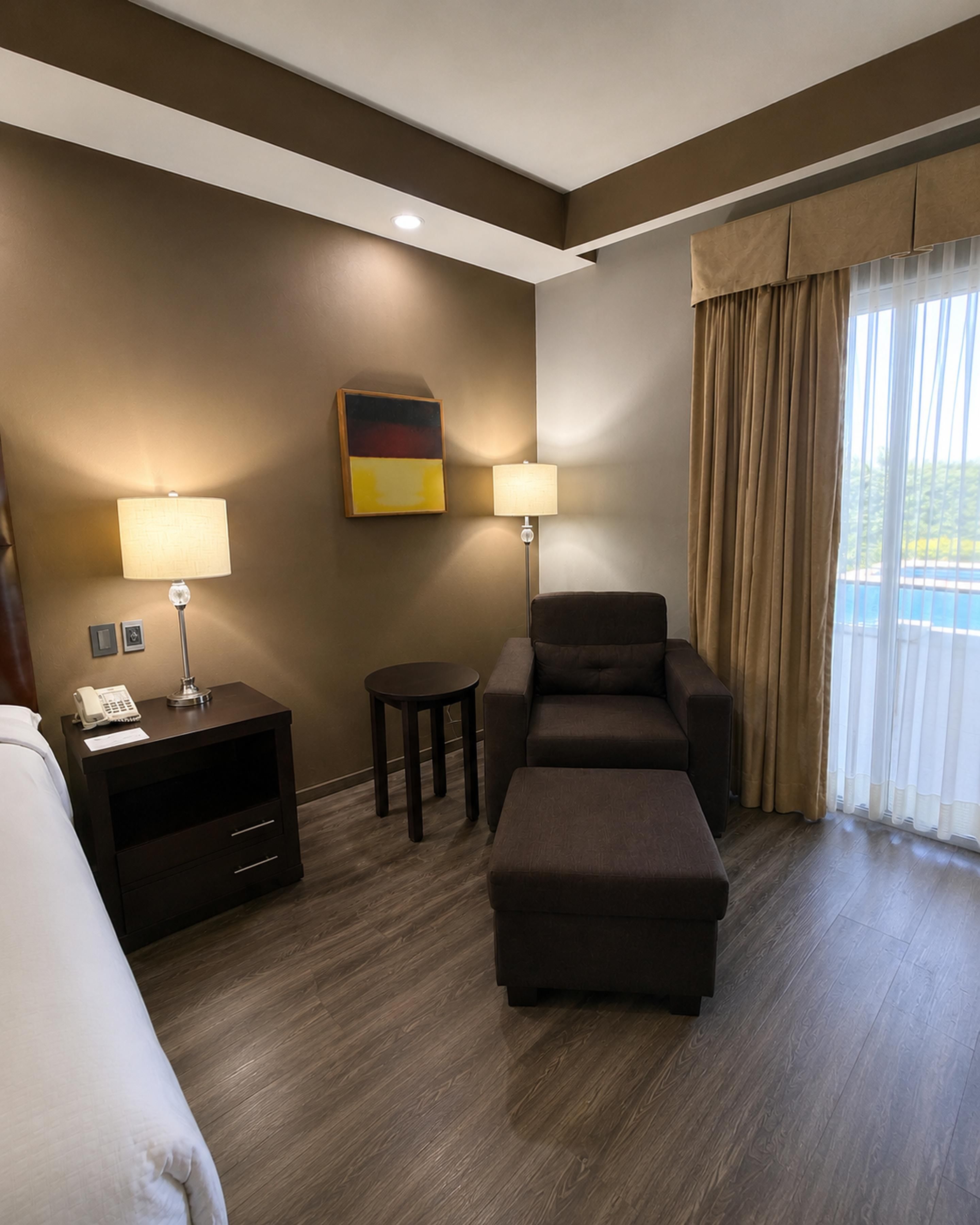 Una habitación de hotel con una cama, un escritorio con una lámpara, un sofá, un reposapiés y un cuadro en la pared.
