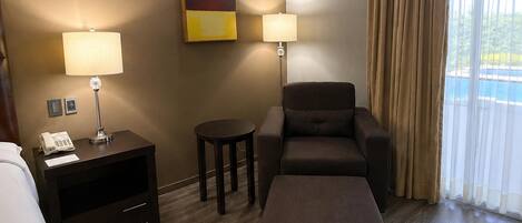 Una habitación de hotel con una cama, un escritorio con una lámpara, un sofá, un reposapiés y un cuadro en la pared.