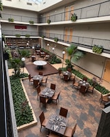 Un área de comedor al aire libre con varios niveles, mesas y sillas, rodeada de plantas en macetas y una mesa redonda central.