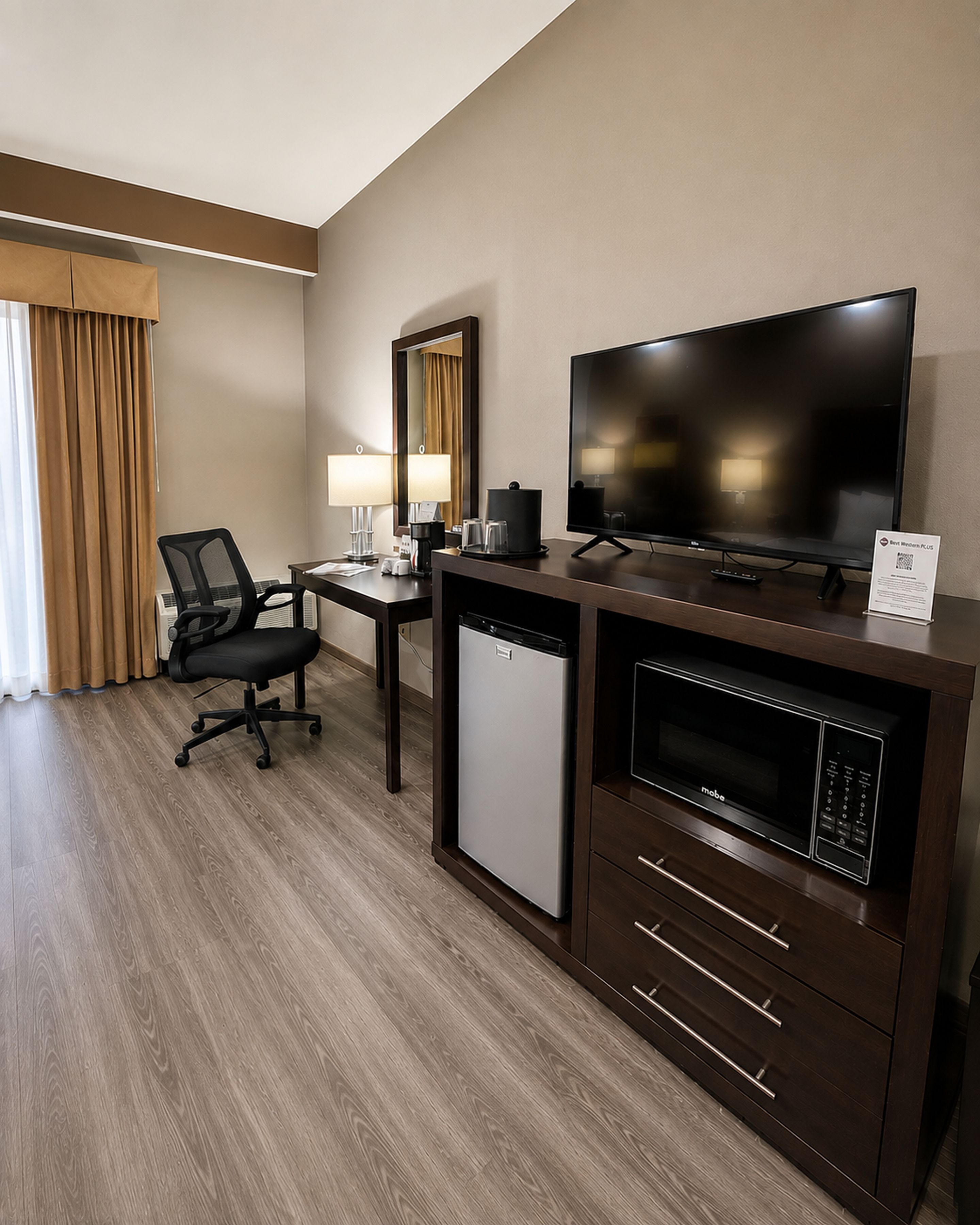 A hotel room with a desk, chair, television, microwave, and a small refrigerator.