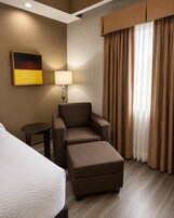 Habitación de hotel con cama, una silla, una mesita, lámpara y ventana con cortinas.