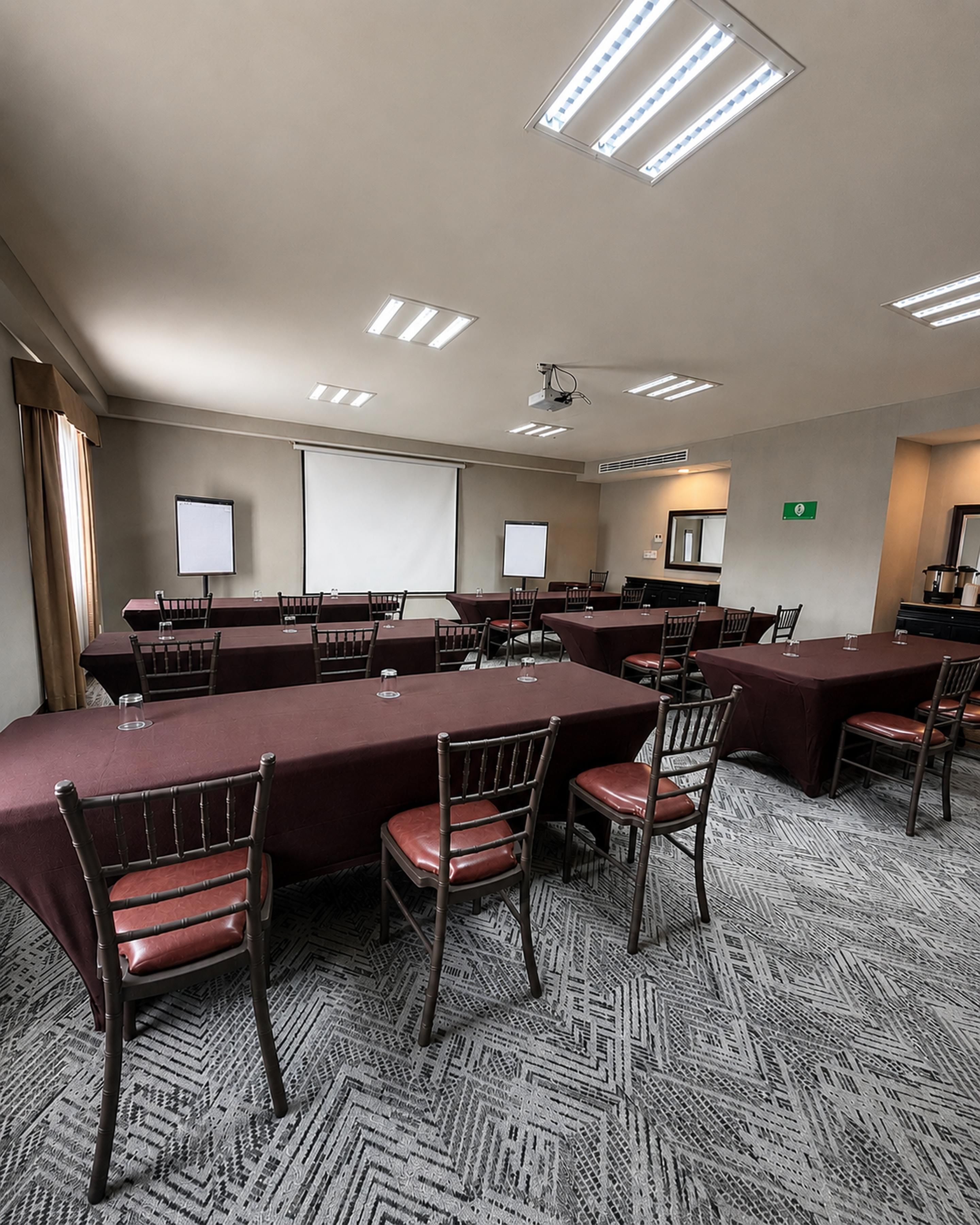 A conference room with multiple tables, chairs, a projector screen, and ceiling lights.