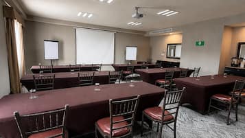 Un salón de conferencias con varias mesas, sillas, una pantalla de proyector y luces en el techo.