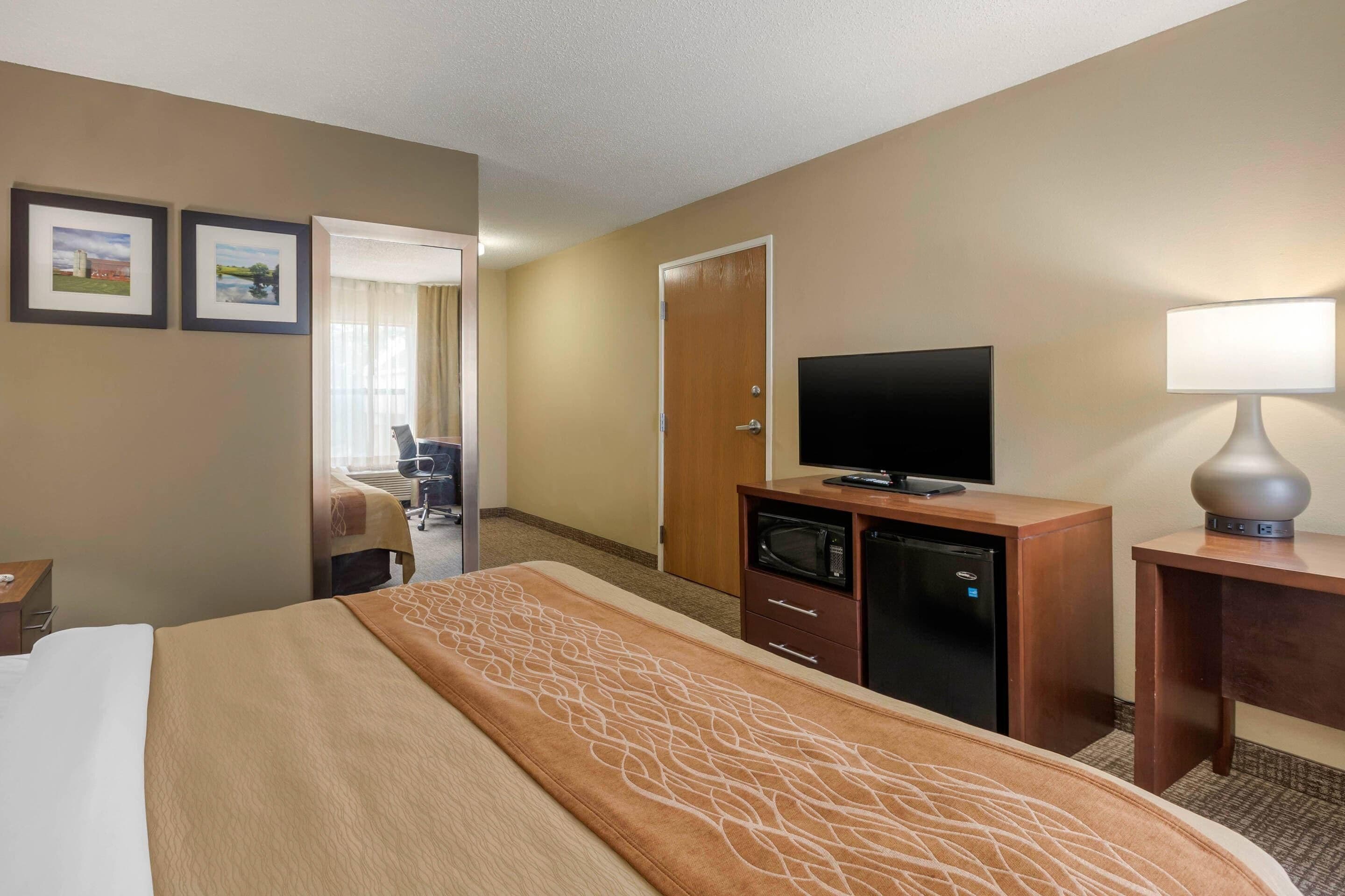 Habitación de hotel con cama, escritorio con televisor, microondas y lámpara. Se ve una puerta que da a otra habitación y una silla en un rincón.