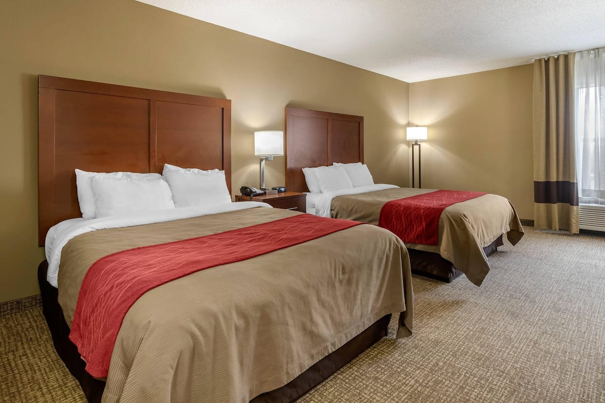 Una habitación de hotel con dos camas, ropa de cama color beige y rojo, cabeceros de madera, lámparas de noche y suelo alfombrado.
