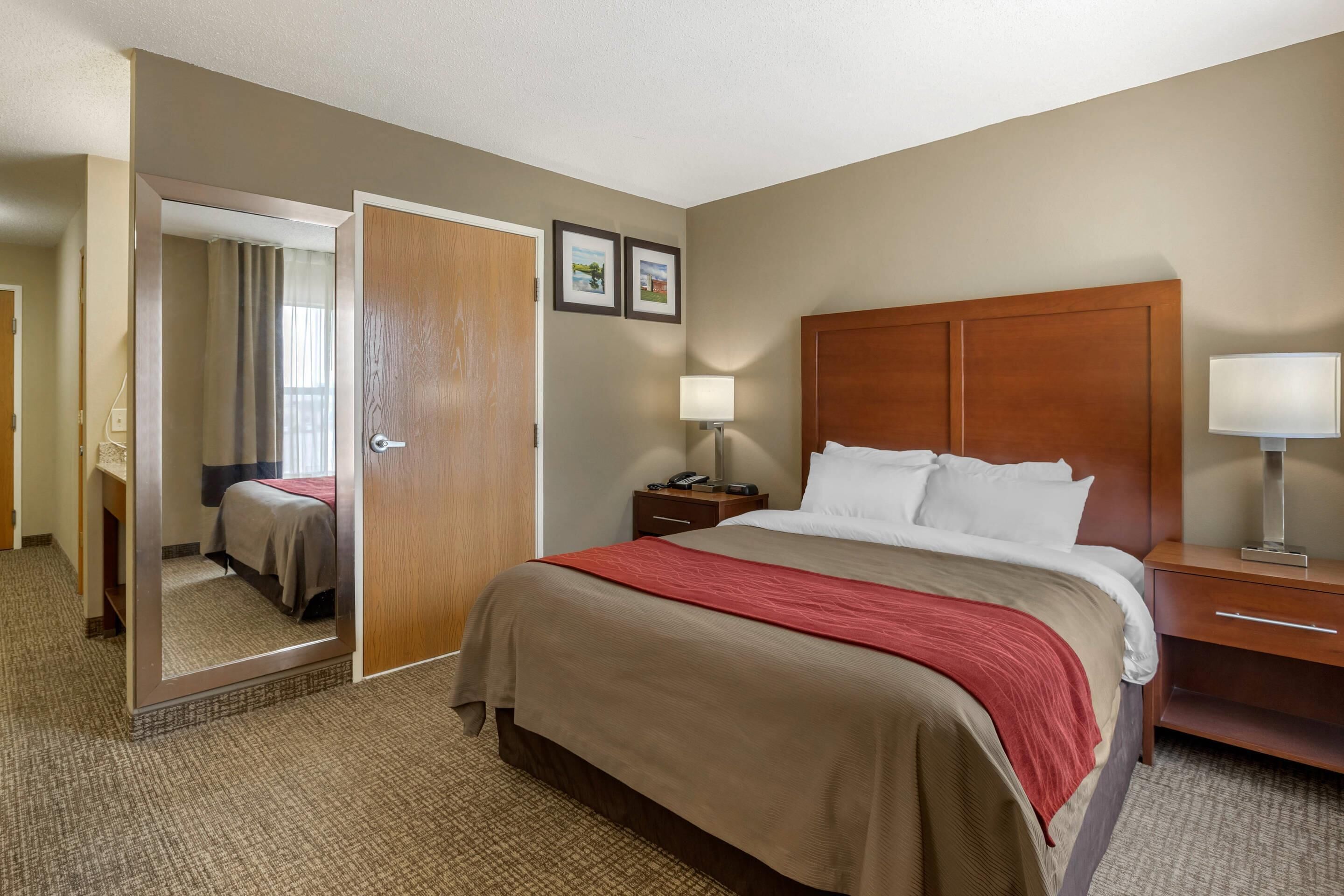 A hotel room with a large bed, a wooden headboard, a nightstand with a lamp, a mirror, and a door leading to a bathroom.