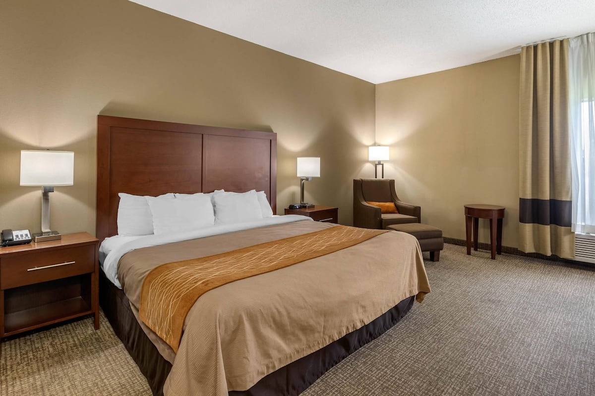 Habitación de hotel con una cama grande, dos mesitas de noche, una silla, una mesita pequeña y una ventana con cortinas.