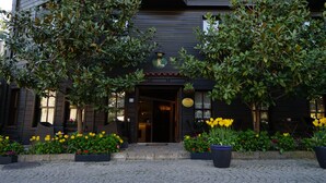 A building with a dark facade, a wooden door, and a sign above the entrance. There are potted plants with yellow flowers in front of the building. - Yusufpasa Konagi - Special Class (Istanbul)