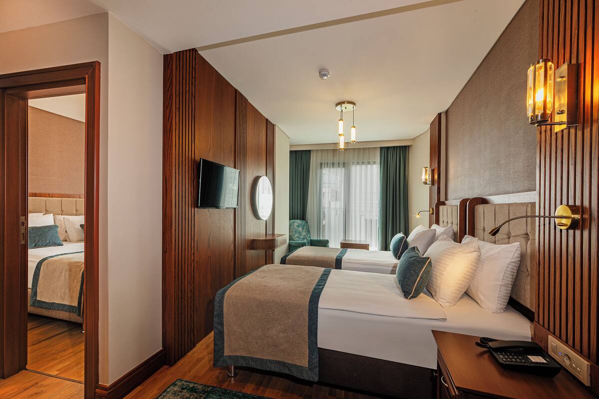 A hotel room with two beds, a television, a desk with a telephone, and a view of another room.