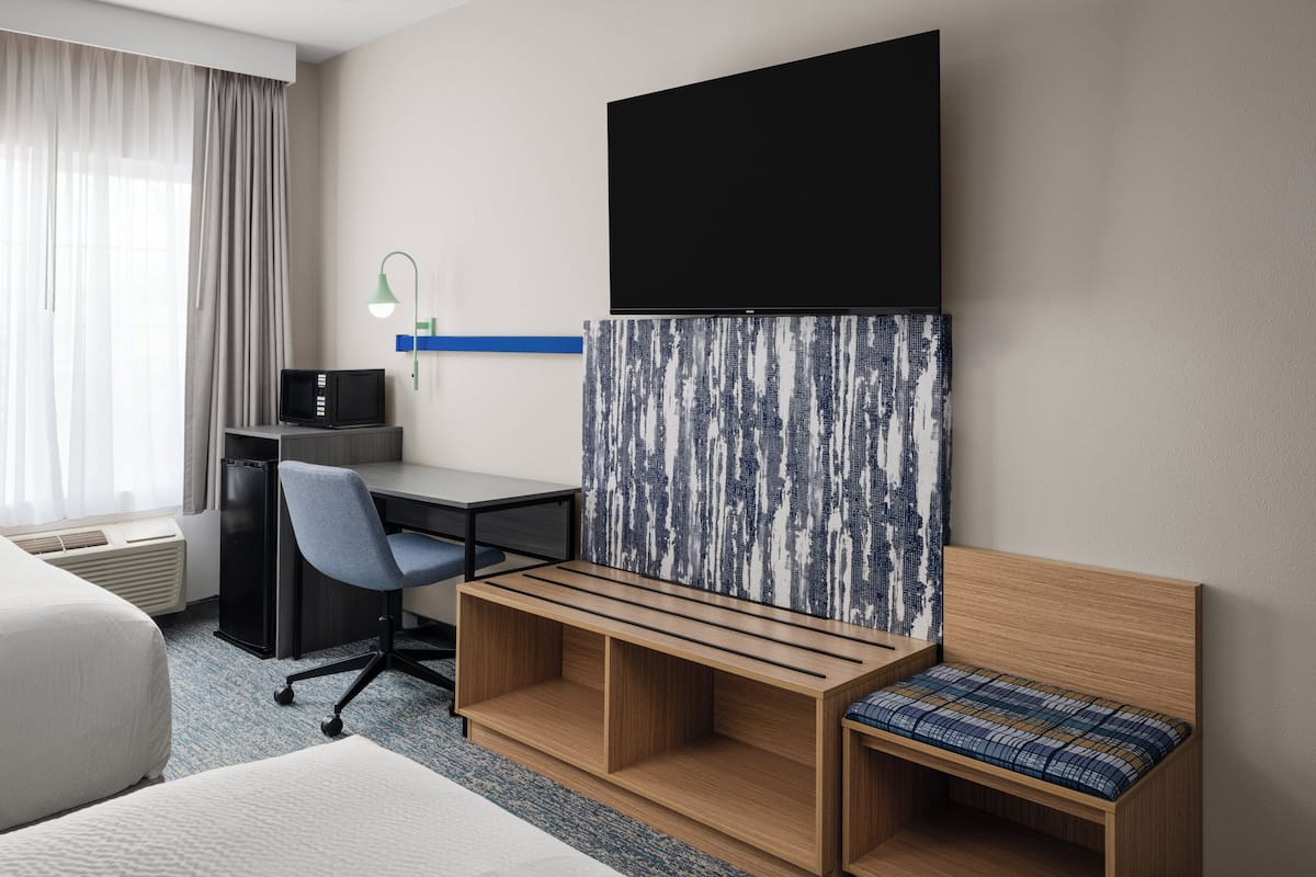Habitación de hotel con cama, escritorio con silla, televisor, refrigerador pequeño y una obra de arte en la pared.