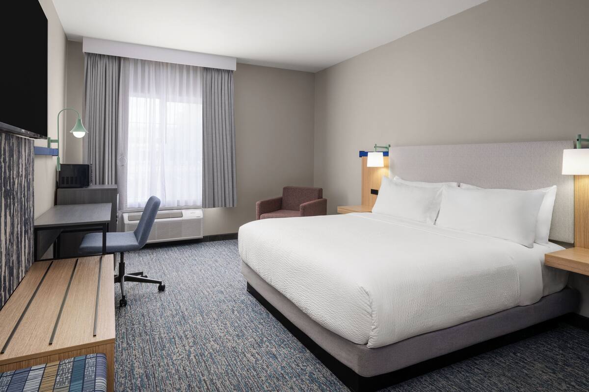 Habitación de hotel con una cama grande, un escritorio con silla, un sillón pequeño, televisión y ventana con cortinas.