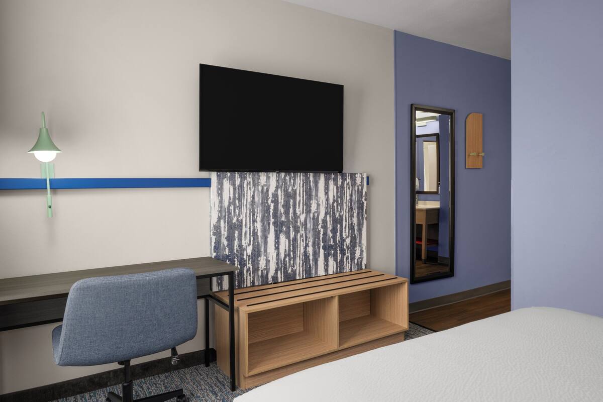 Habitación de hotel con una cama, un escritorio con silla, un televisor montado en la pared, un espejo y un banco de madera.