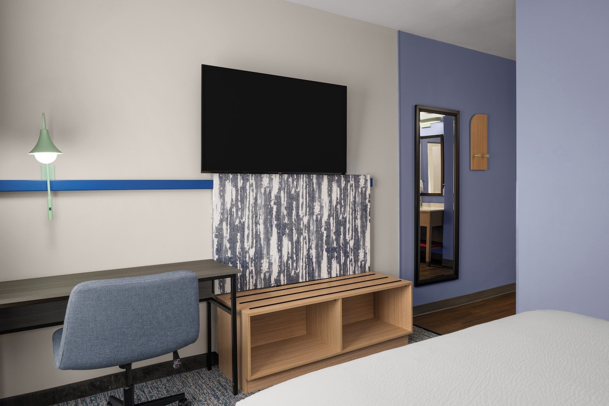 Habitación de hotel con una cama, un escritorio con silla, un televisor montado en la pared, un espejo y un banco de madera.