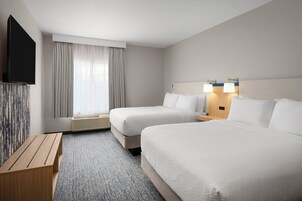 A hotel room with two beds, a flat-screen TV, a wooden bench, and a window with curtains.