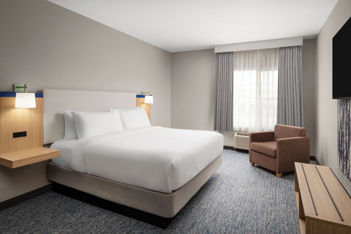 Habitación de hotel con una cama grande, mesitas de noche, una silla, un televisor y una ventana con cortinas.