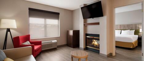 A hotel room with a sofa, a red chair, a round table, a TV, a fireplace, and a bed.