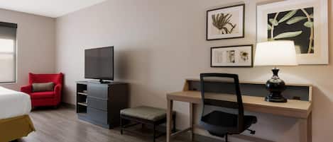 Habitación de hotel con una cama, un sillón rojo, un escritorio con computadora y lámpara, un televisor y obras de arte en la pared.