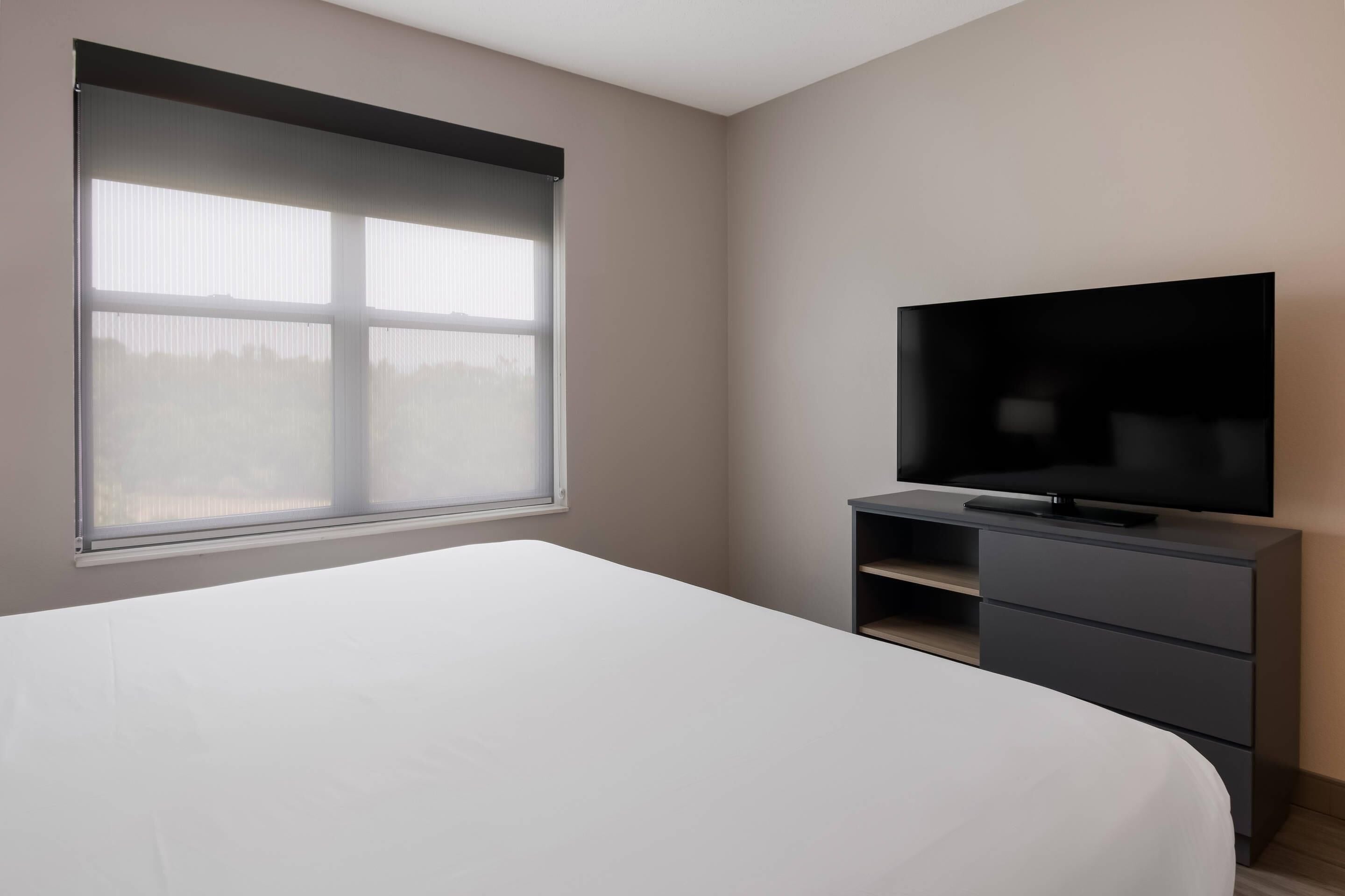 Ein Schlafzimmer mit Bett, einem Fernseher auf einem Schrank, einem Fenster mit Jalousien und einem an der Wand befestigten Regal.