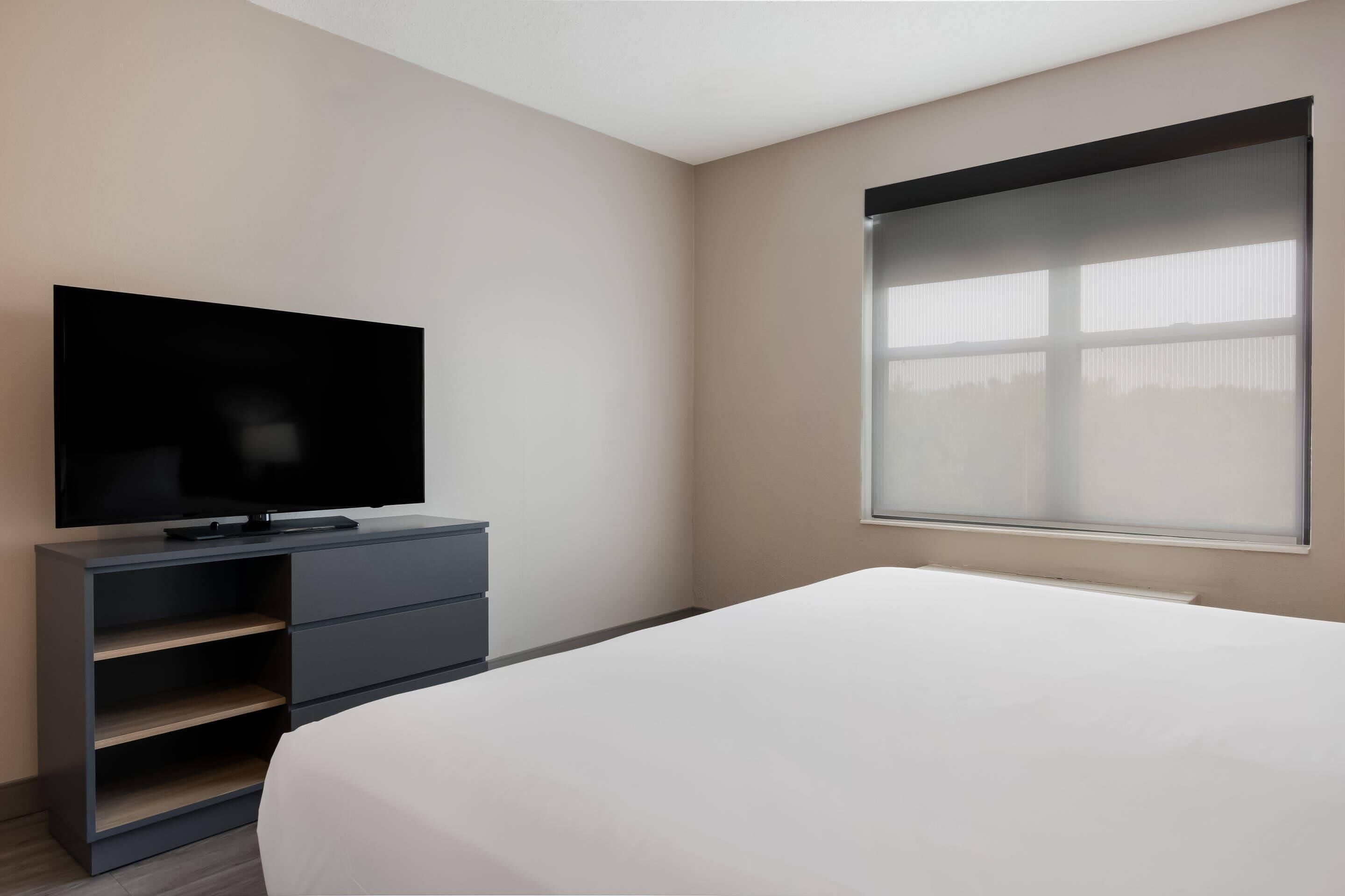 A modern hotel room with a flat-screen TV on a dark dresser, a large bed, and a window with blinds.