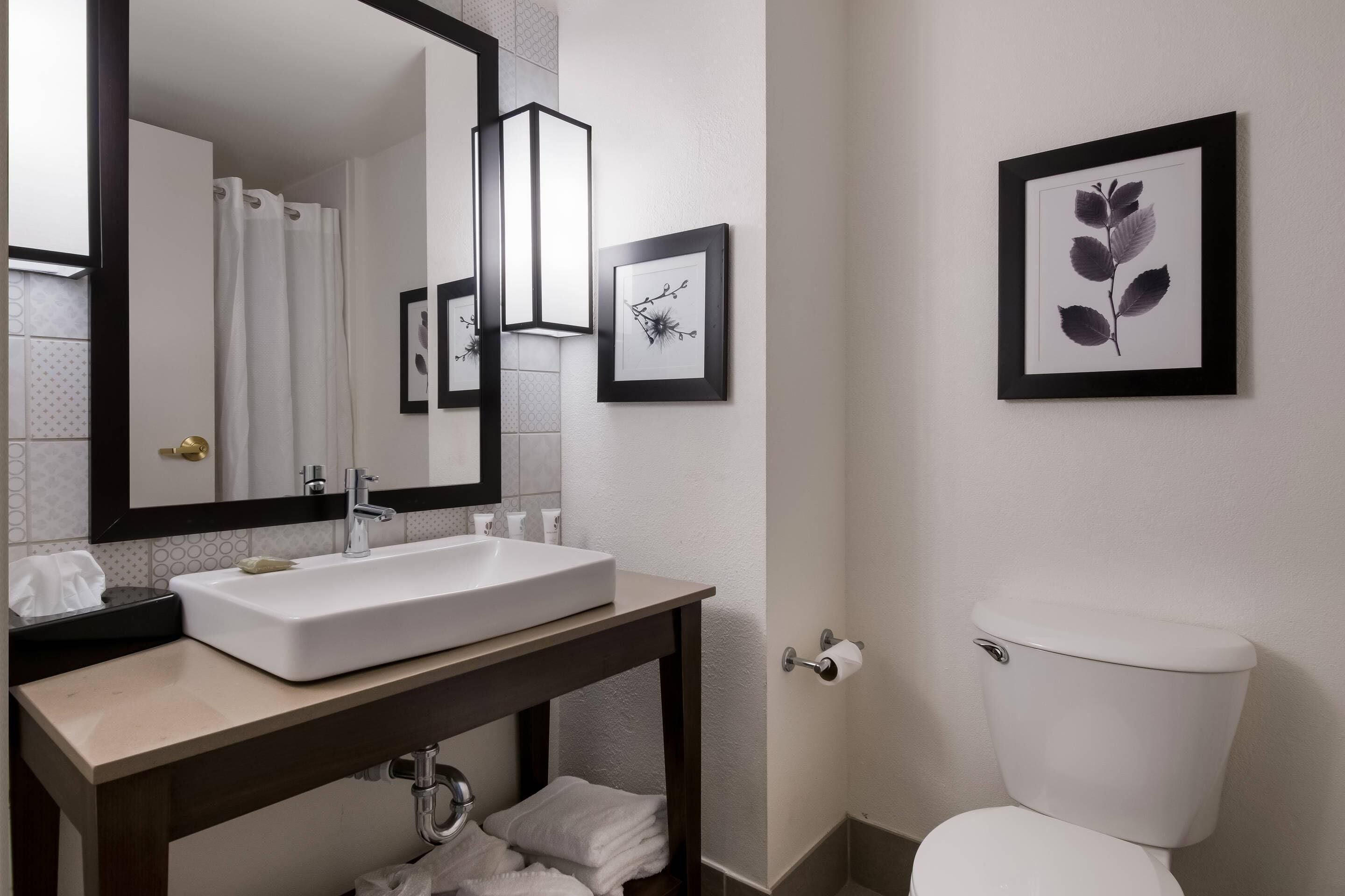 A bathroom with a white sink, mirror, and toilet, framed artwork on the wall, and a towel rack.