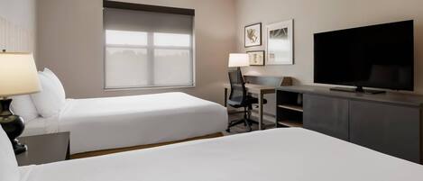 A hotel room with two beds, a desk, a TV, and a window with blinds.