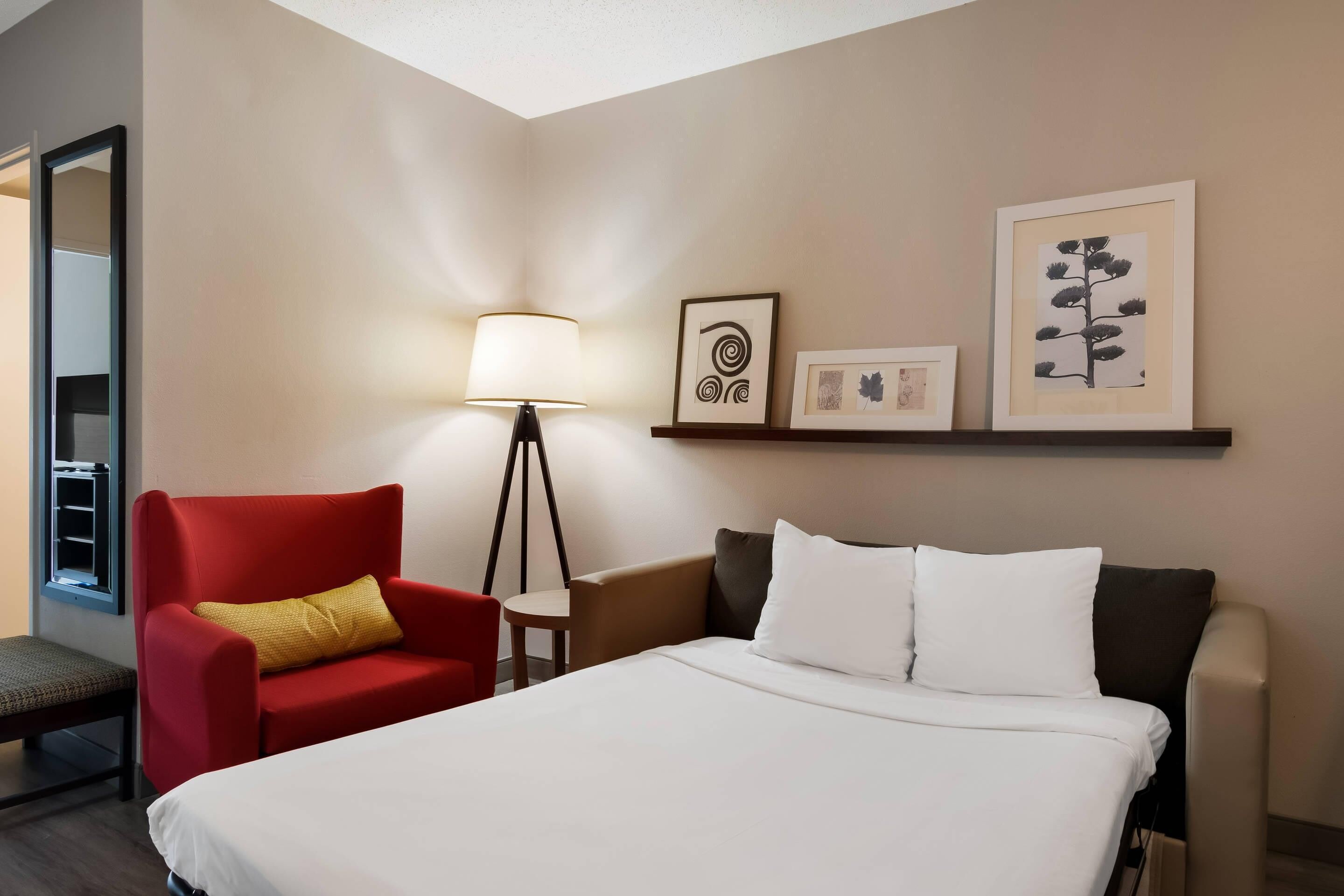 A hotel room with a bed, a red armchair, a small table, a floor lamp, and wall art.