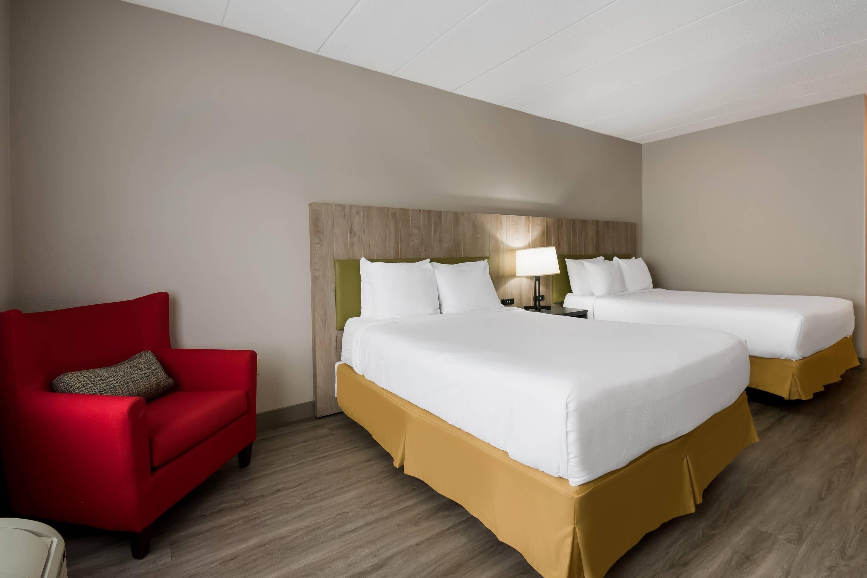 Quarto de hotel com duas camas, uma poltrona vermelha, mesa de cabeceira com abajur e cabeceira de madeira.