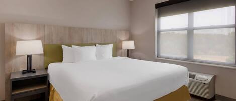 A hotel room with a large bed, two bedside tables with lamps, a window with blinds, and a wall-mounted air conditioner.