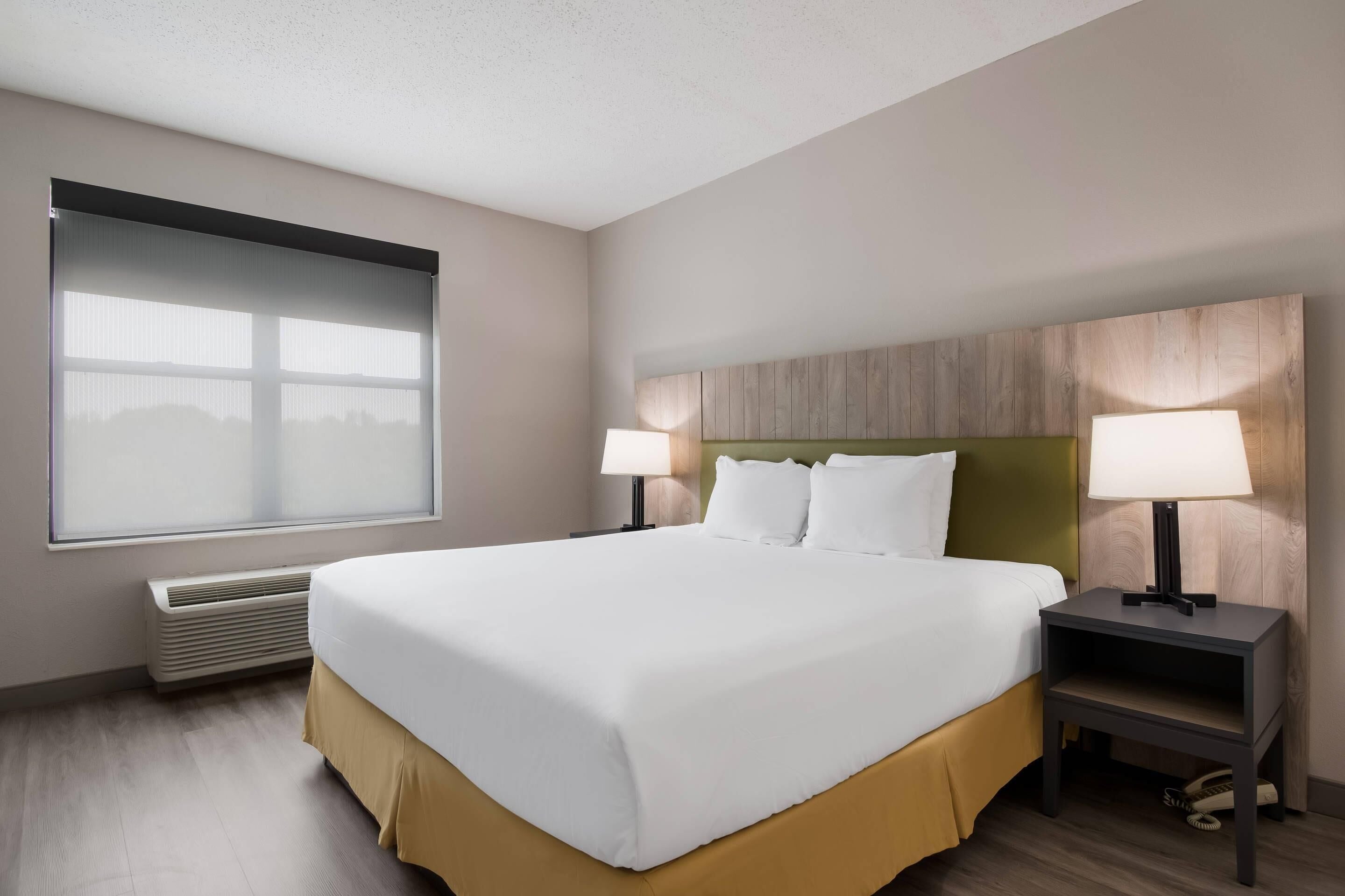 Une chambre d’hôtel avec un grand lit, deux tables de chevet avec des lampes, une fenêtre avec des stores et un radiateur fixé au mur.