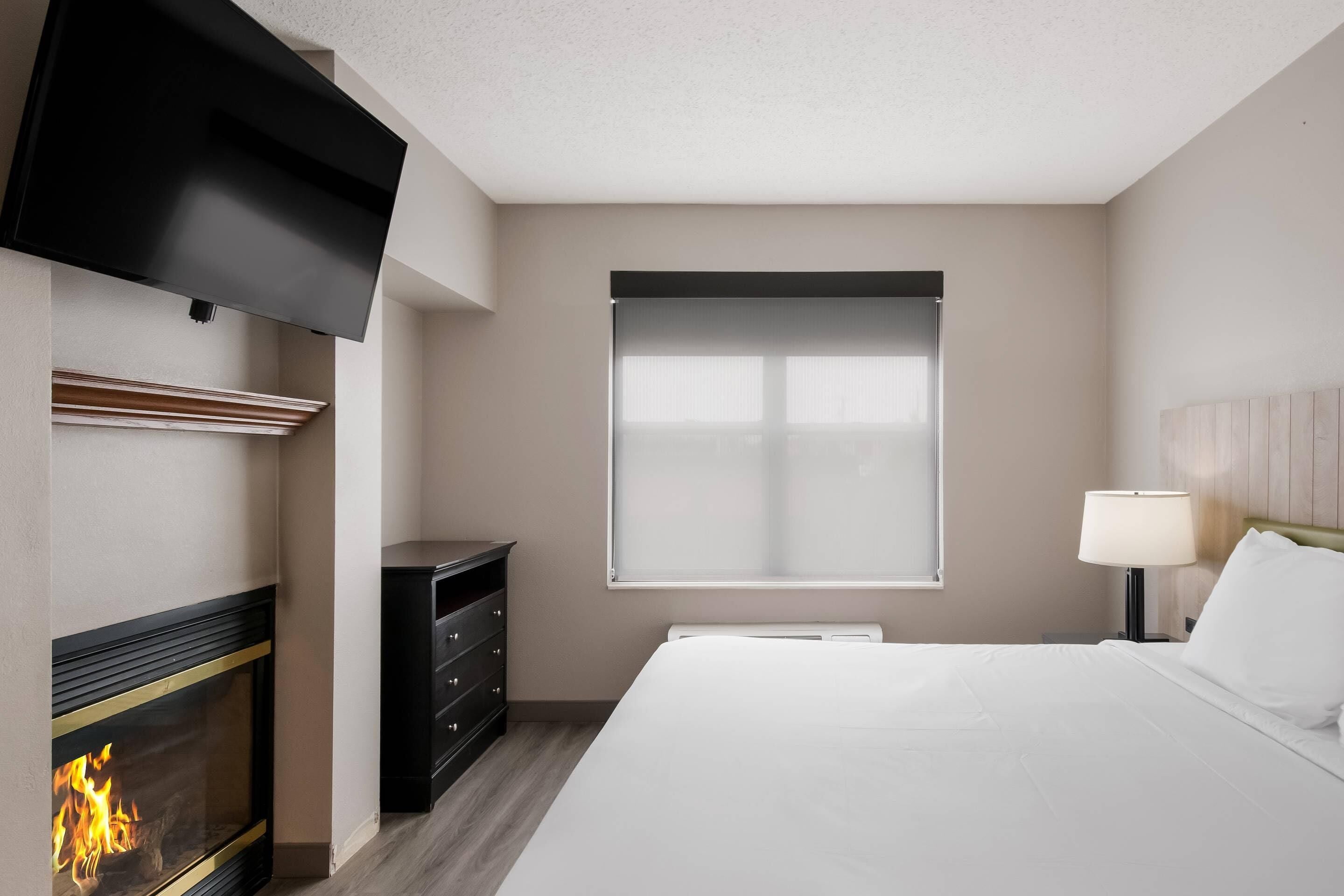 A hotel room with a fireplace, a bed, a TV mounted on the wall, a window with blinds, and a bedside table with a lamp.