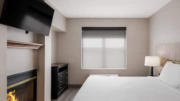 A hotel room with a fireplace, a bed, a TV mounted on the wall, a window with blinds, and a bedside table with a lamp.