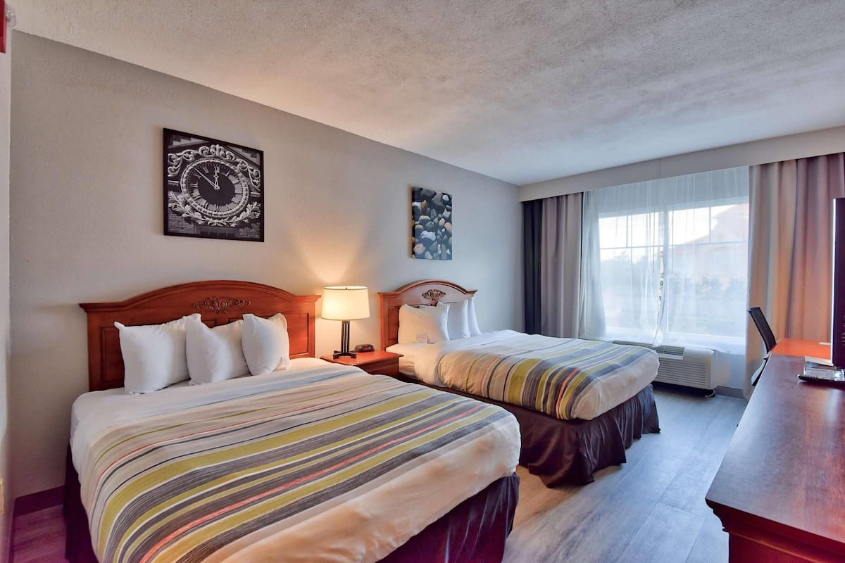 A hotel room with two beds, a wooden headboard, a nightstand with a lamp, a painting on the wall, a window with curtains, and a desk with a laptop.