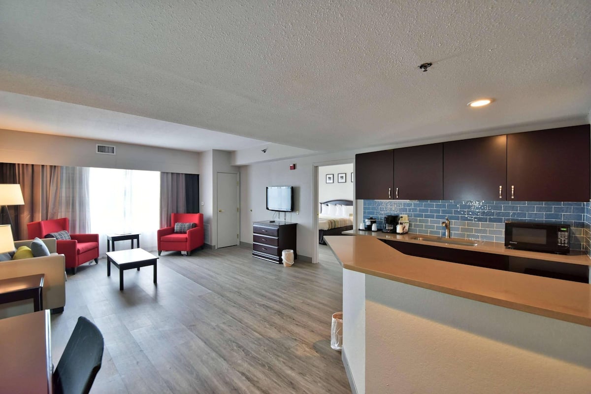 A modern hotel room with a living area, red armchairs, a flat-screen TV, a kitchen with a microwave, and a large window.