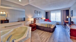 Habitación de hotel con una cama grande, zona de estar con escritorio y silla, televisión, bañera amplia y ventana con cortinas.