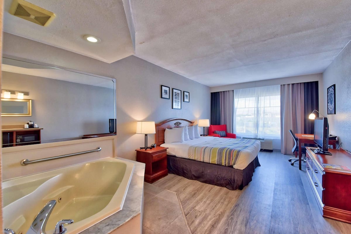 A hotel room with a large bed, a sitting area with a desk and chair, a television, a large bathtub, and a window with curtains.