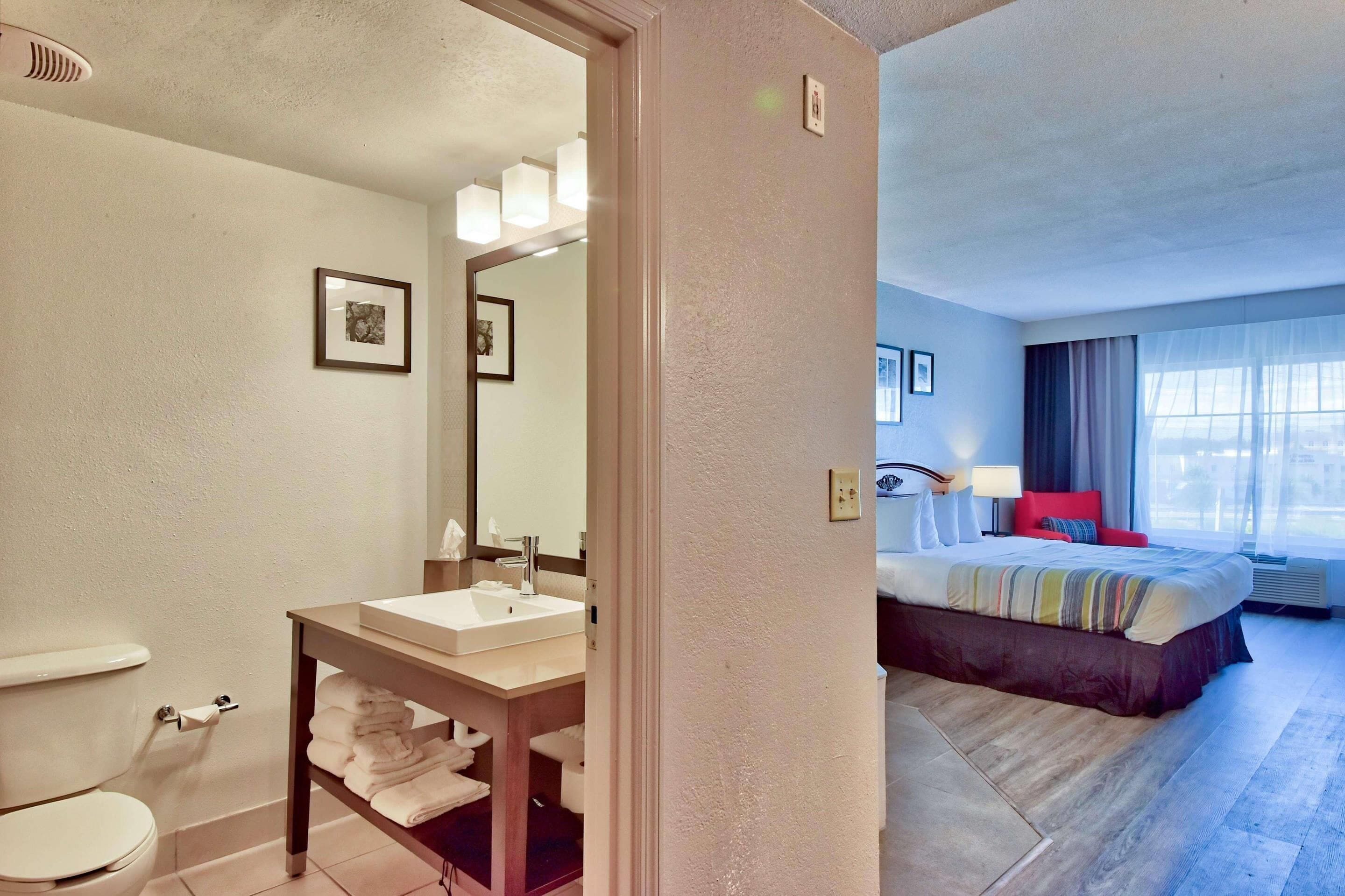 Un baño de hotel con inodoro, lavamanos y perchero. La habitación también tiene una cama con cabecera roja, una mesita de noche y una ventana con cortinas.