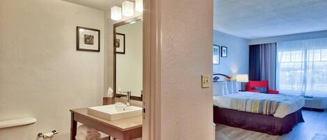 Un baño de hotel con inodoro, lavamanos y perchero. La habitación también tiene una cama con cabecera roja, una mesita de noche y una ventana con cortinas.